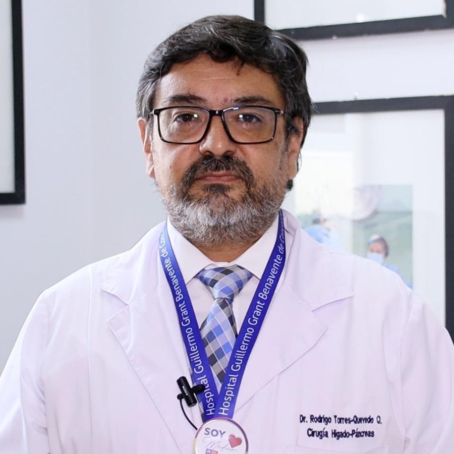 Male doctor with glasses and a gray beard in a white lab coat, blue striped tie, and a blue hospital lanyard wearing a microphone mic.