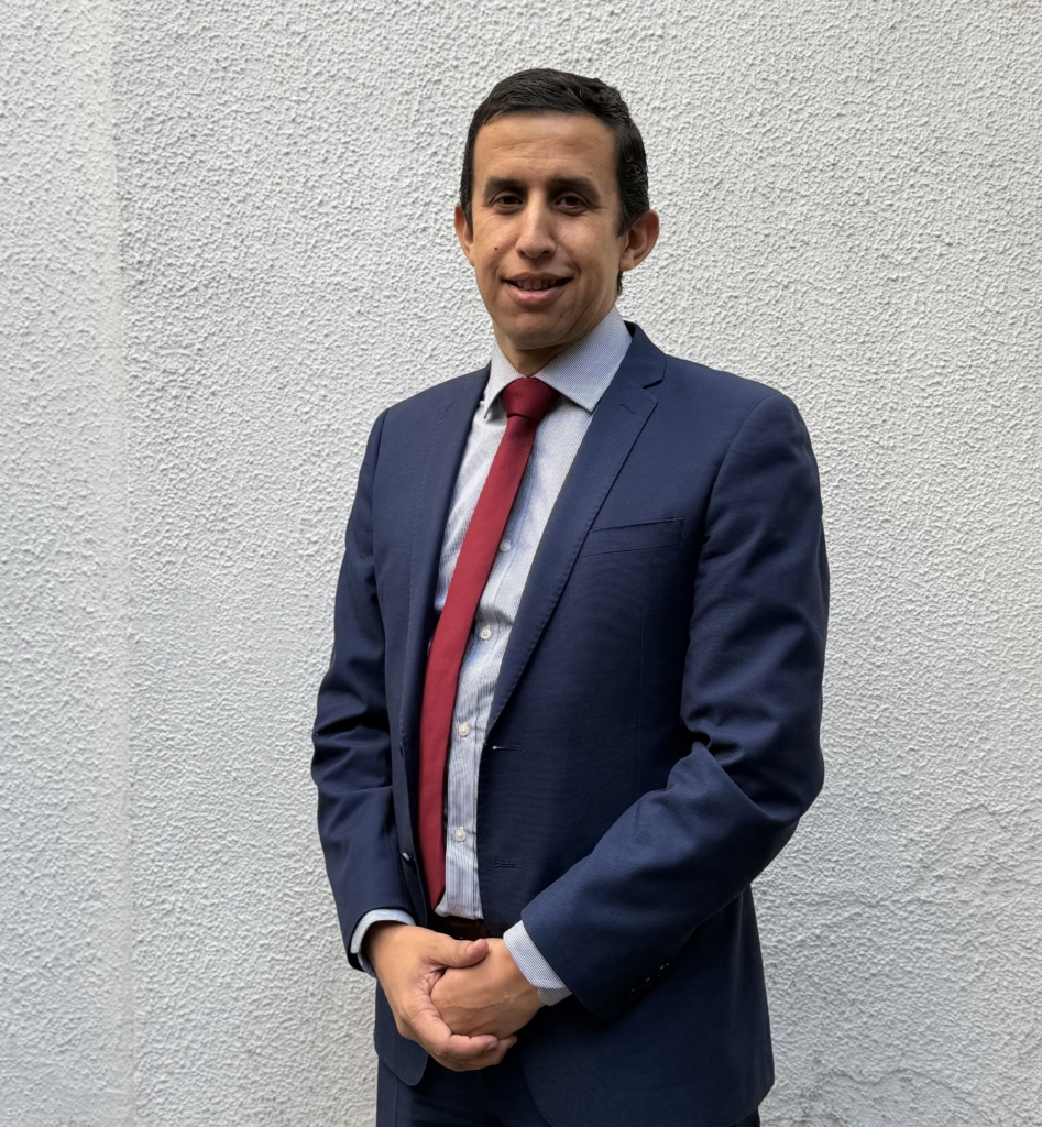 Man in a navy suit and red tie stands against a textured gray wall, hands clasped in front and a subtle smile.