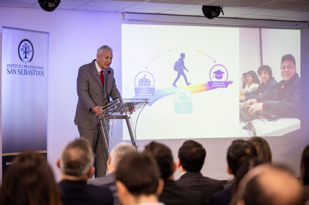 Captura-de-pantalla-2026-05-18-144908 - Instituto Profesional San Sebastián Speaker in a gray suit delivers a talk at a podium, with a projected infographic and students on computers behind him in a conference room.