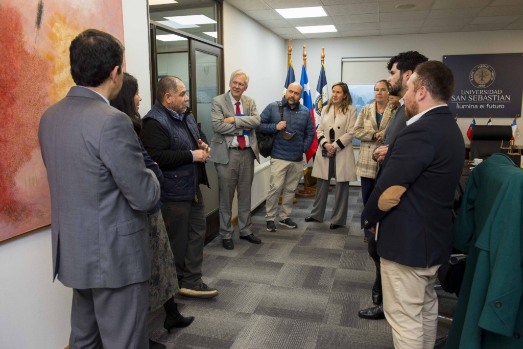 0001 - Instituto Profesional San Sebastián Group of professionals in a university office hallway talking in a circle, with flags and a Universidad San Sebastián sign in the background, signifying a formal meeting.