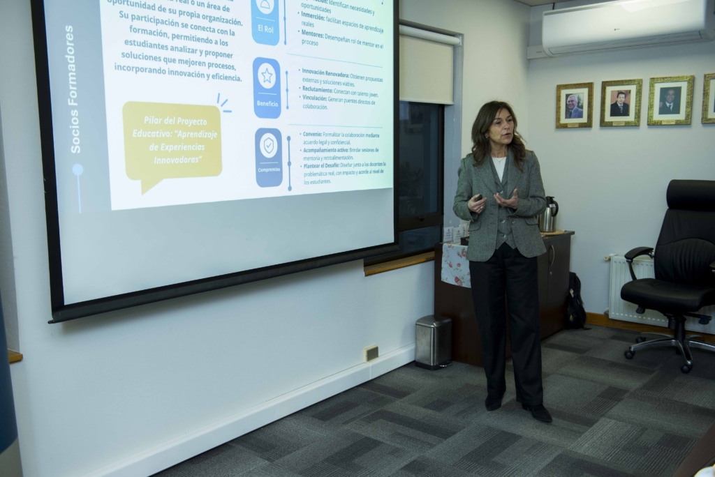 0028 - Instituto Profesional San Sebastián Presenter standing beside a projected slide in a classroom, gesturing as she speaks; wall portraits line the back of the room.