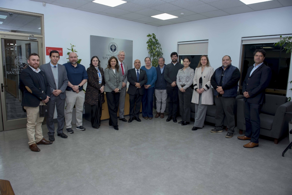 0044 - Instituto Profesional San Sebastián Group of professionals in business attire posing in a modern office lobby with a signage plaque and plants behind them.