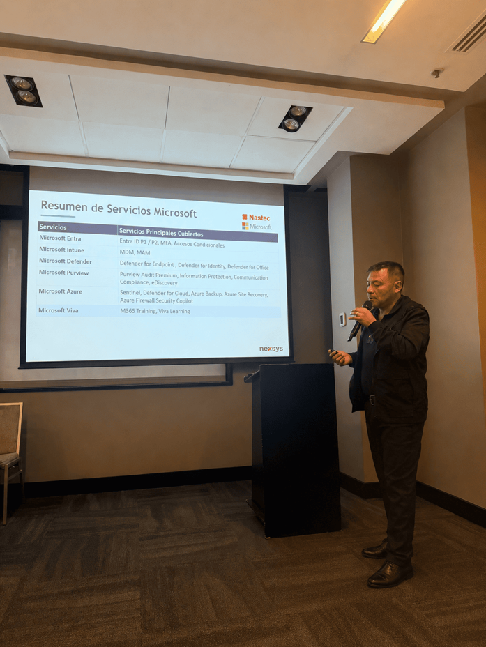 Man giving a presentation in a conference room, standing at a black podium beside a projector screen displaying a slide titled 'Resumen de Servicios Microsoft'.