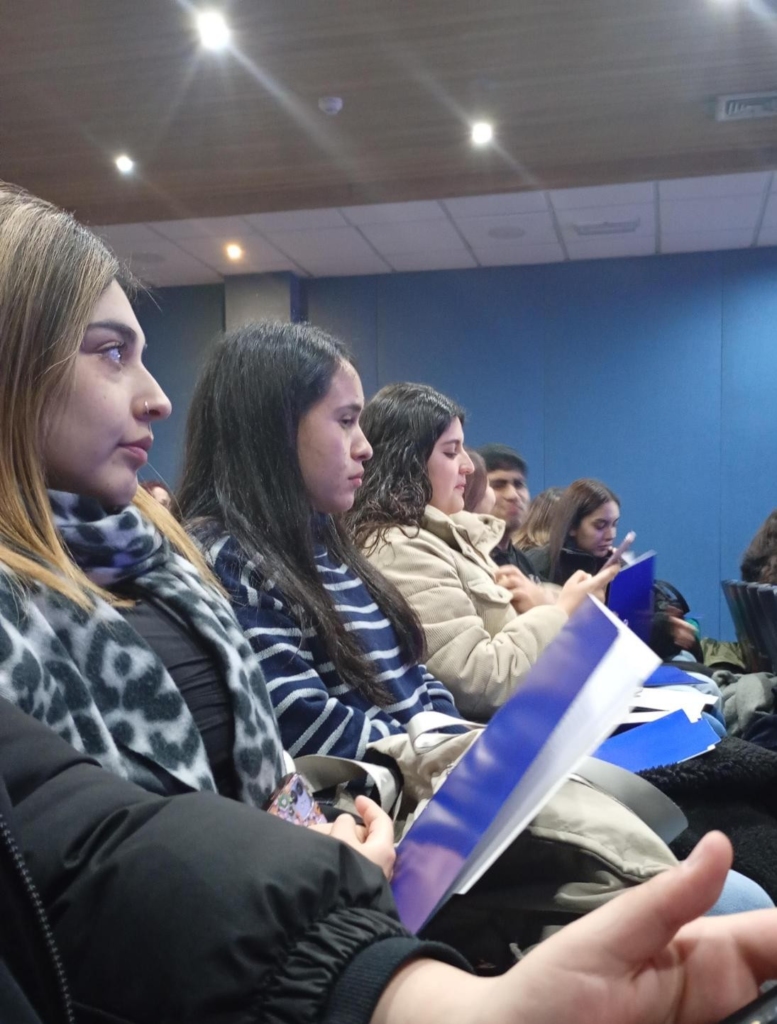 Group of people sitting in a row at a lecture, each holding blue-covered folders or programs, focused on the event.