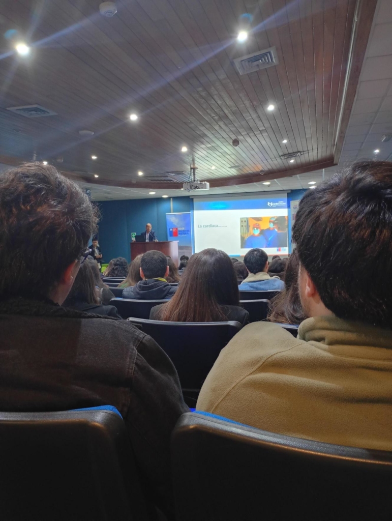 Lecturer at a podium addressing an audience in a lecture hall with a projected slide on the screen behind him.