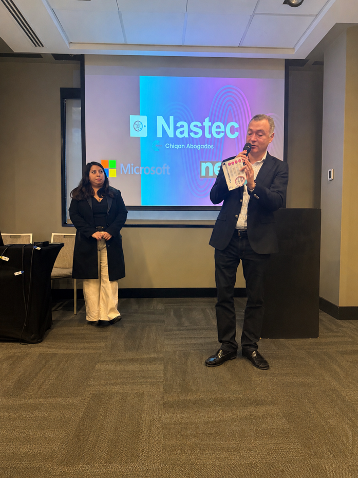 Man in a dark suit speaks into a handheld microphone, holding papers, as a woman in a black coat stands nearby in a conference room; a presentation screen behind shows company logos including Nastec and Microsoft.