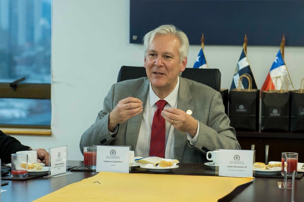 Image 35 - Instituto Profesional San Sebastián Older man in a gray suit and red tie speaks at a conference table with nameplates and snacks in front of him.