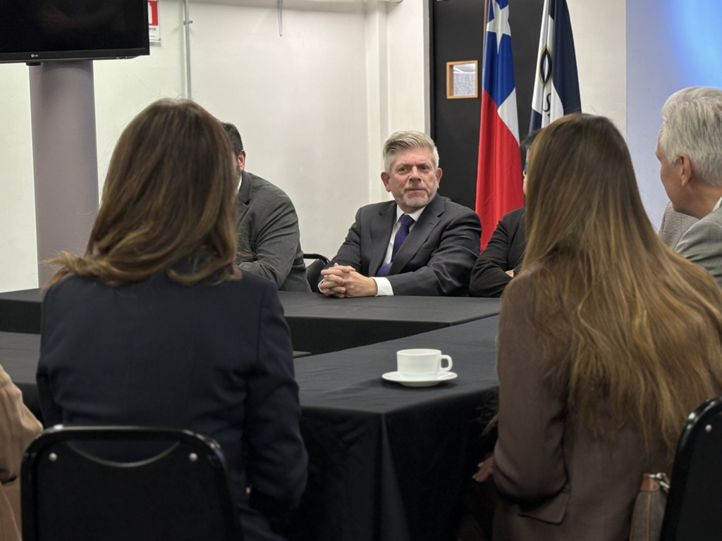 Man in a dark suit and purple tie sits at a conference table, facing a group of people in a formal meeting room with flags in the background.
