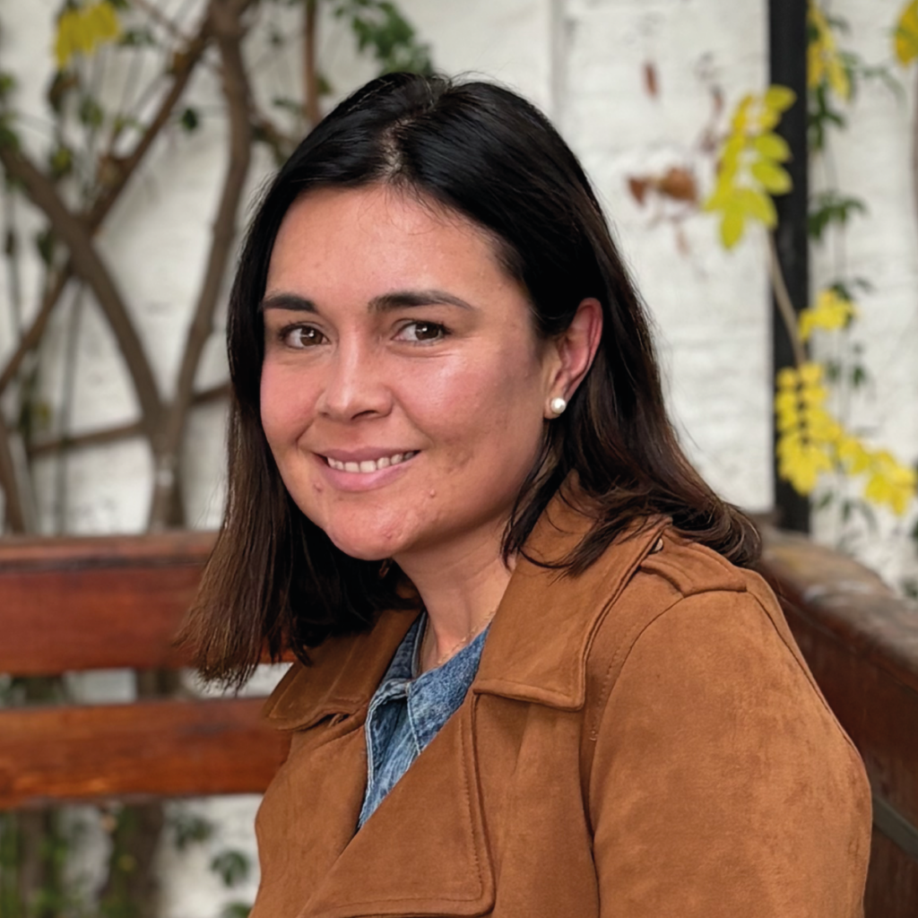 Fotografía-1200-x-1200 1 - Instituto Profesional San Sebastián Smiling woman with shoulder-length dark hair and pearl earrings, wearing a brown suede jacket, outdoors beside a wooden bench.