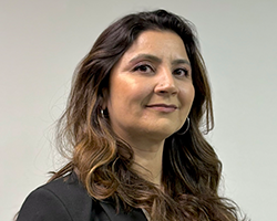 Portrait of a woman with long wavy brown hair, wearing a dark blazer, looking slightly to the left against a neutral background.